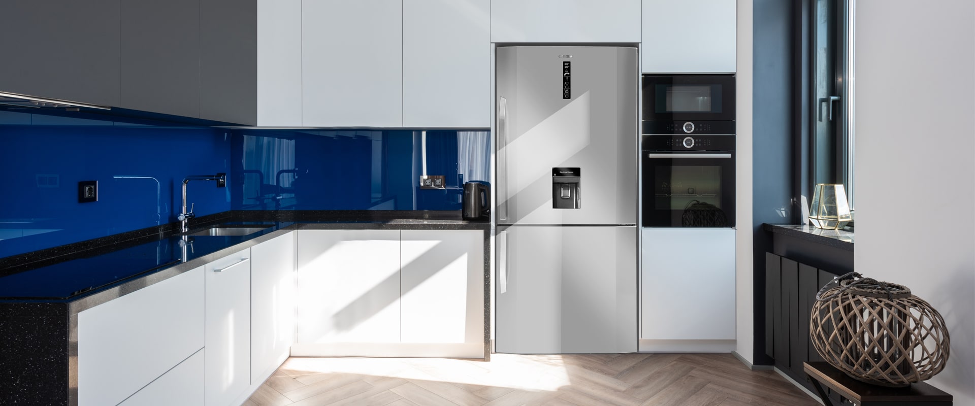 Modern kitchen with white cabinets, blue backsplash, black countertops, stainless steel refrigerator, and built-in ovens.