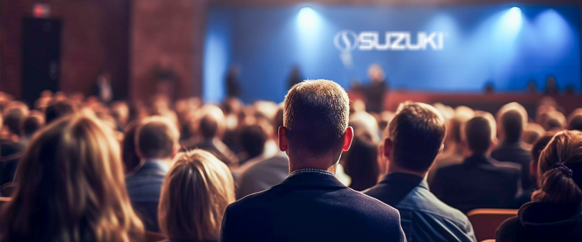 Back view of an audience attending a Suzuki event with the company logo illuminated on a large blue screen on stage.
