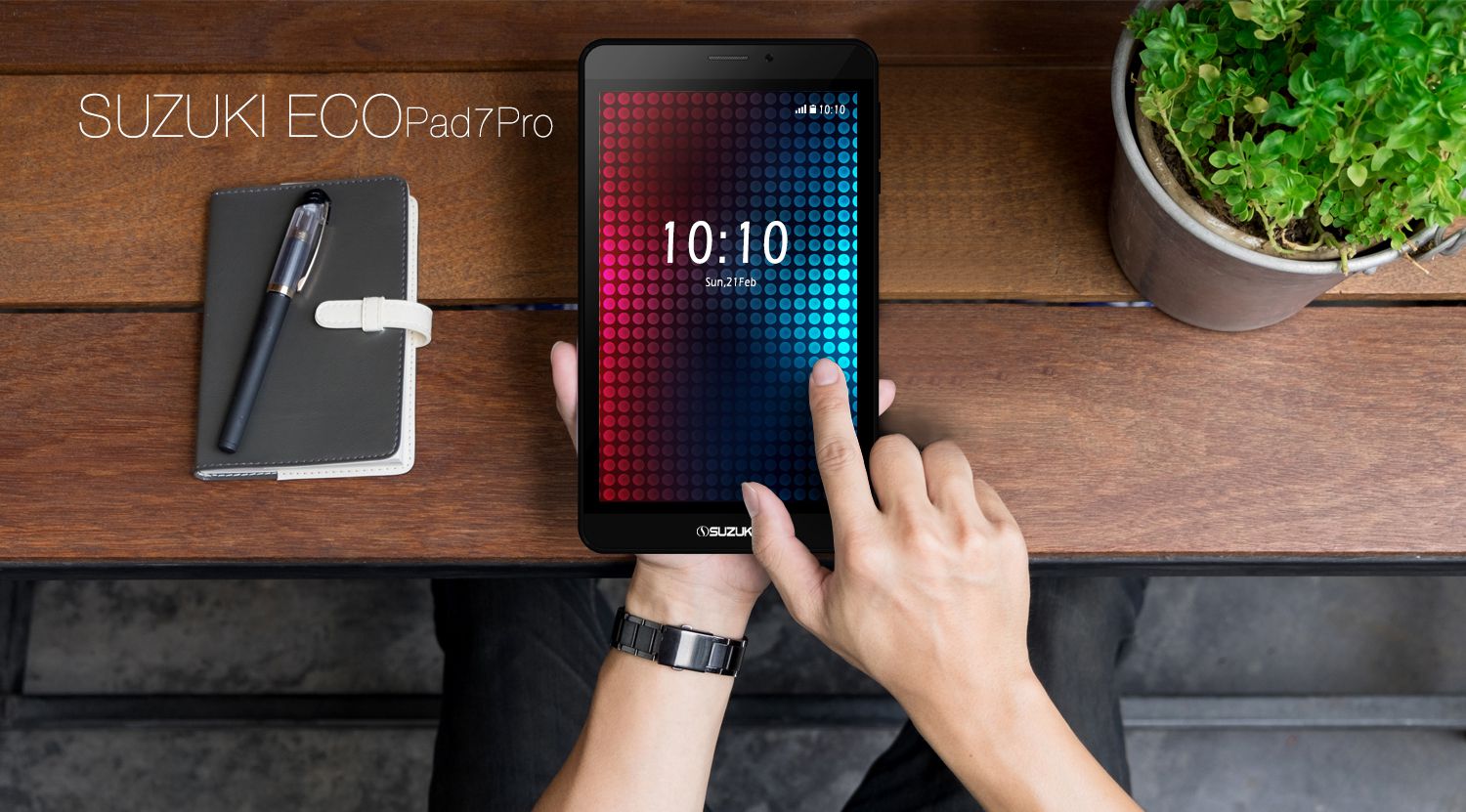 Hands holding the Suzuki ECO Pad 7 Pro tablet showing the lock screen on a wooden table with a notebook and potted plant.