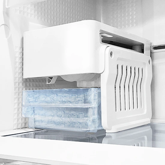 A white automatic ice maker installed inside a freezer with two clear bins of ice cubes stacked below it.